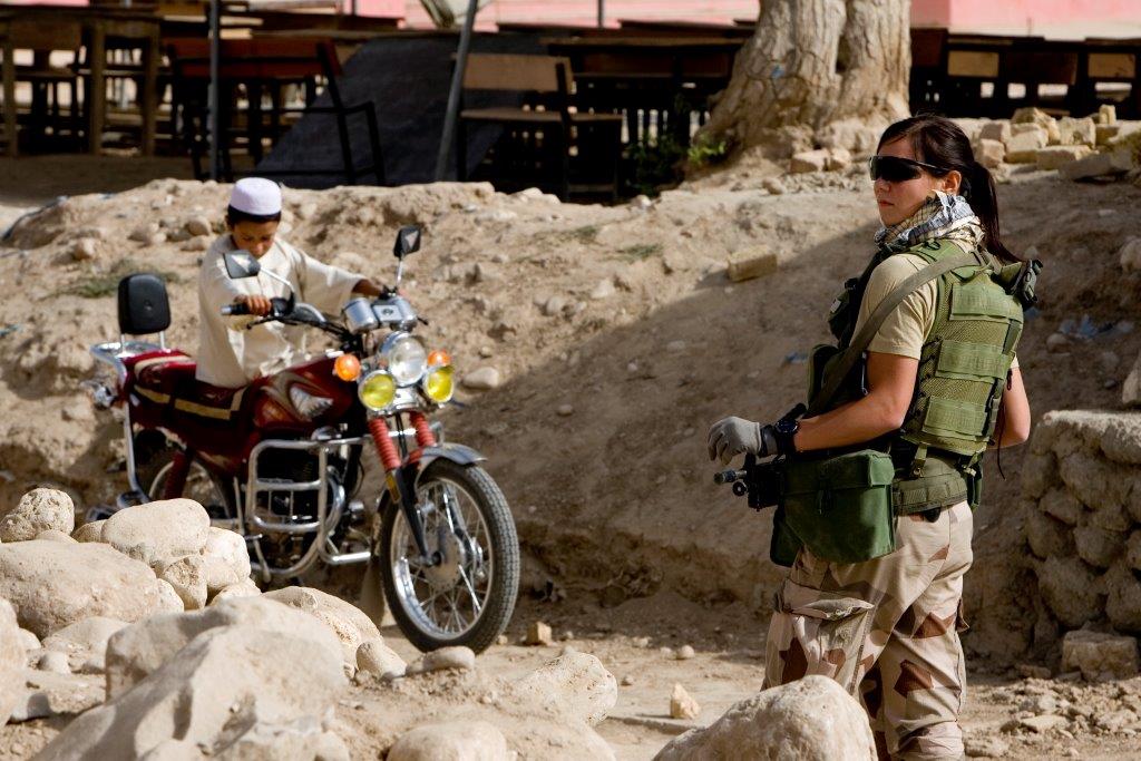 Kvinnlig militär i solglasögon framför afghansk pojke på röd motorcykel.